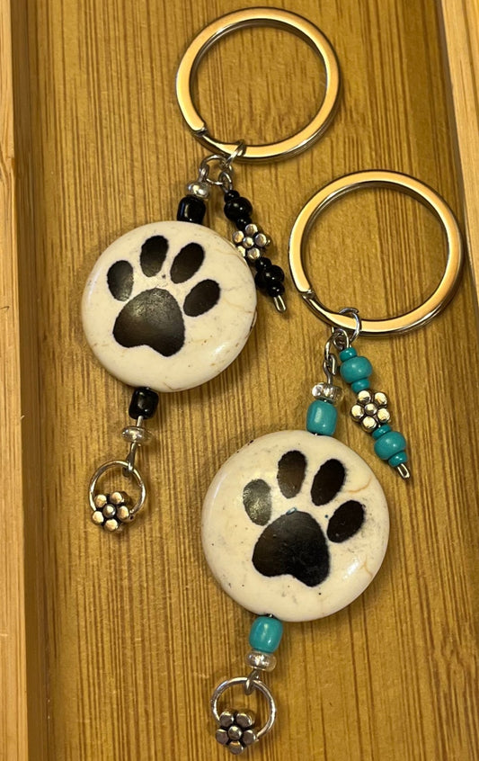 Two keychains, each has a ceramic beige coin bead with black paw prints on either side, accented with two metal flower charms and either black or turquoise glass beads.
