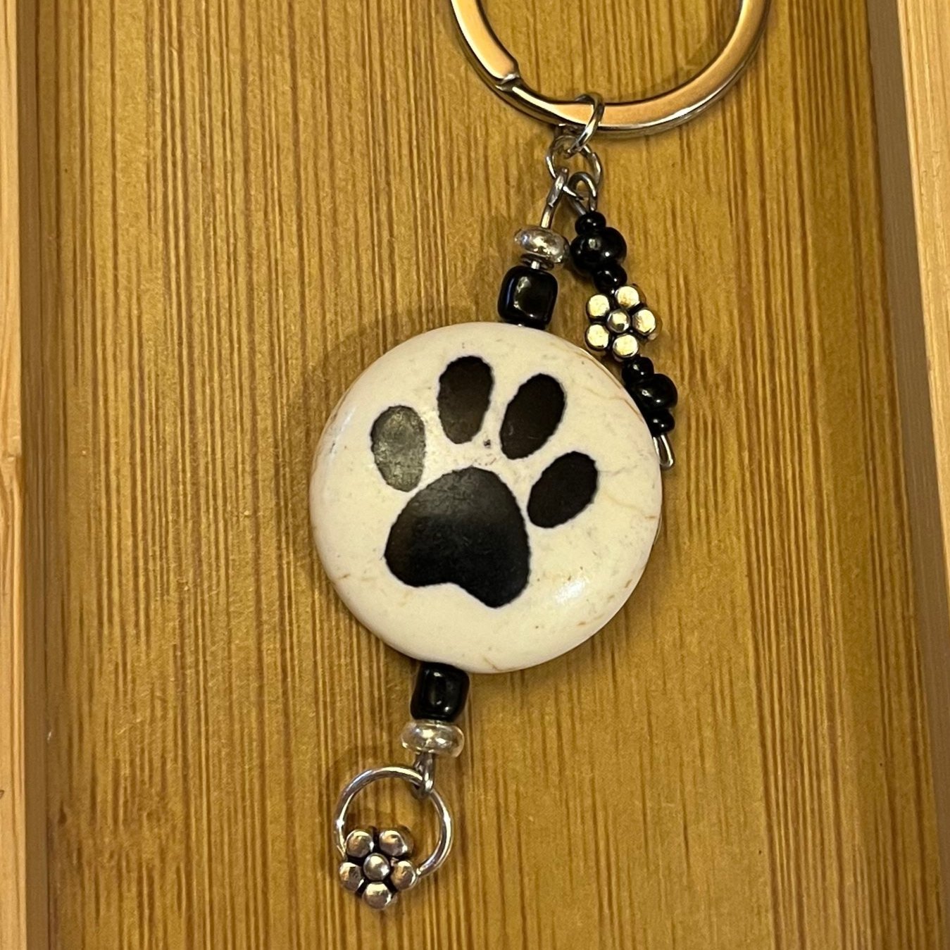 Keychain with a ceramic beige coin bead with black paw prints on either side, accented with two metal flower charms and either black glass beads.