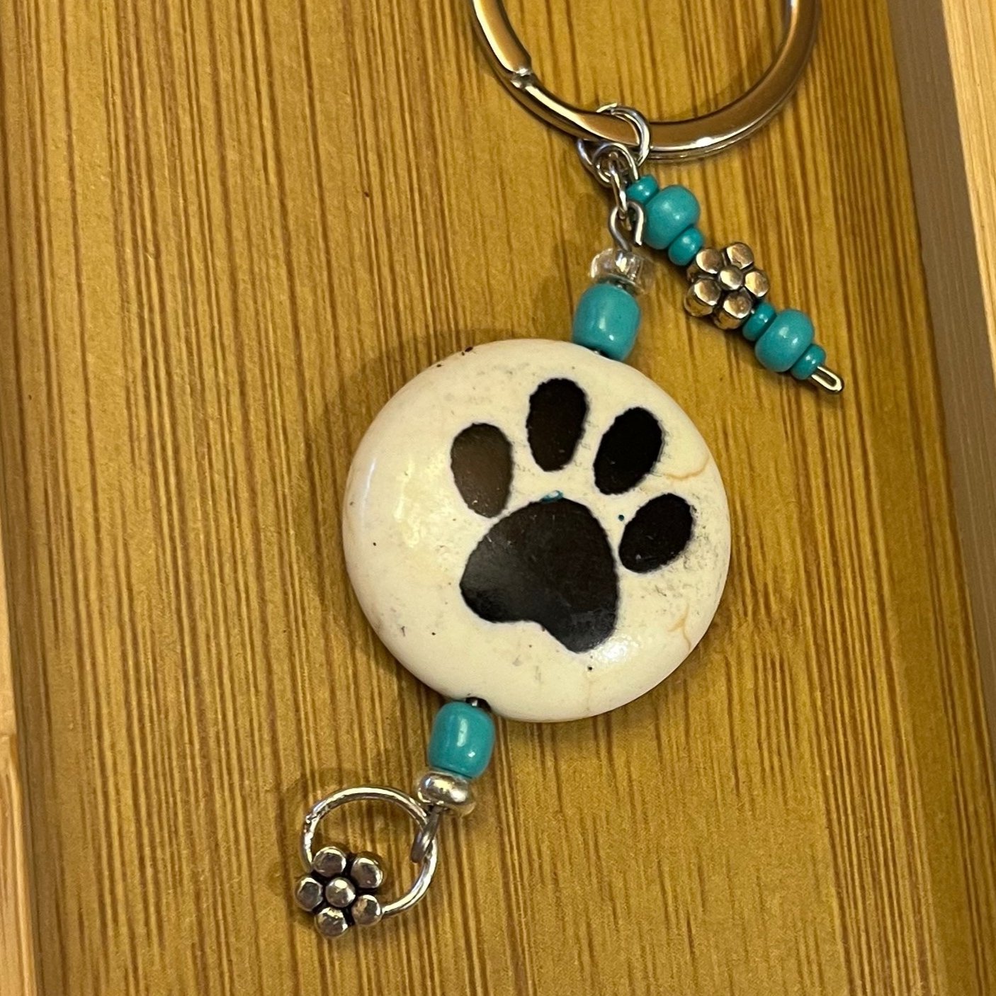 Keychain with a ceramic beige coin bead with black paw prints on either side, accented with two metal flower charms and turquoise glass beads.