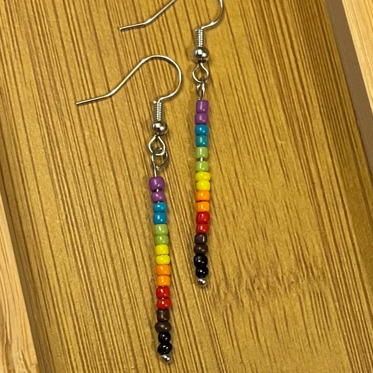 Pride stick earrings with the progressive rainbow colours in opaque glass seed beads: purple, blue, green, yellow, orange, red, brown, and black.